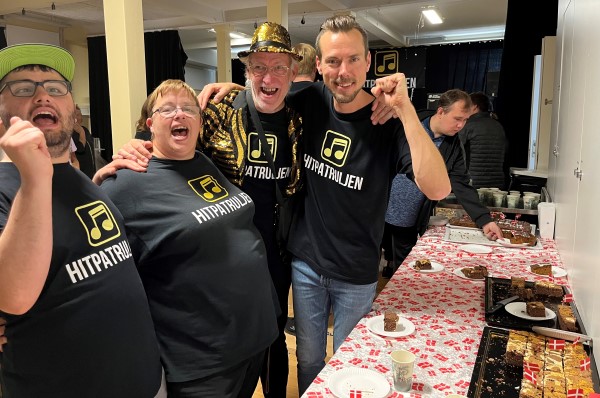 De glade bandmedlemmer iført Hitpatruljen-tshirt ved kagebordet med flag.